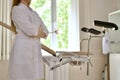 A doctor in white uniform with folded hands. Stands in profile in front of a gynecological chair with footrests.