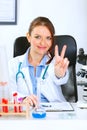 Doctor woman sitting at table and showing victory Royalty Free Stock Photo