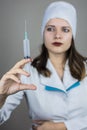 Doctor in white coat with dark hair in medical blue mask holds syringe in hands on light background