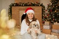 Dog sitting by christmas tree with its owner, charming woman with toothy smile sitting on floor in festive living room, woman Royalty Free Stock Photo