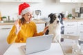 Dog and woman wearing Santa hat waving hand video calling family by webcam. Girl with laptop having virtual meeting chat on Royalty Free Stock Photo