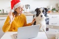 Dog and woman wearing Santa hat waving hand video calling family by webcam. Girl with laptop having virtual meeting chat on Royalty Free Stock Photo