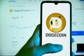 The Dogecoin cryptocurrency logo is seen on the display of a mobile phone with a laptop with a stock chart in the background in Ba Royalty Free Stock Photo