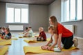 Doing butterfly exercise. Young female trainer helping little girl to stretch her legs. Group of children sitting on the