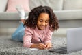 Doing homework. Happy african american girl studying online from home taking notes in front of laptop, lying on carpet Royalty Free Stock Photo