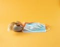 A doll lies down and sleeps on a grey pillow covered with a mask on yellow background. Minimal covid 19 concept