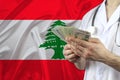 Dollar bills in hands of doctor in uniform on the background of silk national flag of Lebanon, concept of influenza, virus, COVID- Royalty Free Stock Photo