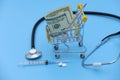 Dollars banknote and thermometer in shopping cart, white pills, syringe and stethoscope on blue background. Medicine concept with Royalty Free Stock Photo