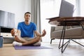 Domestic yoga. Arab man meditating in front of laptop at home, sitting with closed eyes in lotus position