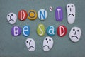 Don`t be sad, creative slogan composed with multi colored stone letters and stone sad faces over green sand Royalty Free Stock Photo