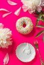 Donut coated with white frosting and colorful sprinkles on crimson background with peony petals, vintage saucer and fork