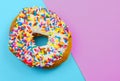 A donut, or doughnut, with white icing and colorful sprinkles on a bright blue and pink geometric background