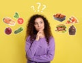 Doubtful African American woman choosing between healthy and unhealthy food on yellow background Royalty Free Stock Photo