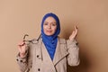 Doubtful Muslim woman in blue hijab, looking at camera, posing with stylish eyeglasses over isolated beige background