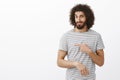 Doubtful unimpressed handsome Eastern guy with beard and curly hairstyle in striped t-shirt, pointing left and lifting