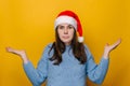 Doubting young girl shrug shoulders, looks uncertain, confused and looking to hand palms, wears Christmas hat and blue sweater Royalty Free Stock Photo