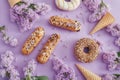 Doughnuts and cream cakes lie on the table among lilac flowers on purple paper. Spring and sweets, delicious cakes. Still life and Royalty Free Stock Photo