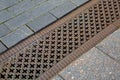 A drainage ditch covered with an iron patterned lattice on a sidewalk made of granite tiles.