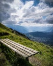 Dramatic alpine panoramic top view across valley with mountains and village in background, wooden bench close up