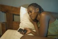 Dramatic lifestyle portrait of young sad and depressed black afro American woman crying in bed holding mobile phone victim of