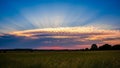 Dramatic sunset sky with radiant sun rays bursting through scattered clouds over a vast green field and distant trees Royalty Free Stock Photo