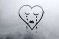 drawn sad face in shape of heart with closed eyes and tears on foggy glass window raindrops blue color concept photo