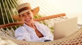 Dream work. Successful female freelancer wearing straw hat using laptop and smiling at camera while lying in the hammock on the Royalty Free Stock Photo