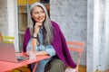 Dreamful senior Asian woman sits at table with computer and drink on cafe terrace