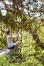 Dreaming beautiful girl sitting on a stone and making selfie on a smartphone surrounded by coniferous forest on a sunny day