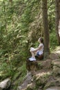 Dreaming beautiful girl sitting on a stone surrounded by coniferous forest on a sunny day. Vertical frame Royalty Free Stock Photo