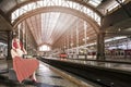 Dreaming and Waiting Caucasian Blond Girl in Pin-up Style Sitting on Suitcase On Railway Station With Trains on Tracks