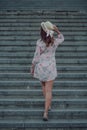 Dreaming young woman in summer dress holding hat and going up on stairways, view from her back Royalty Free Stock Photo