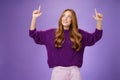 Dreamy girl with red hair and freckles in warm cozy purple sweater raising hands looking and pointing up with intrigued