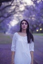 Dreamy image of beautiful woman in white dress walking in street surrounded by purple Jacaranda trees