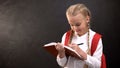 Dreamy little schoolgirl writing in notebook, doing homework against blackboard