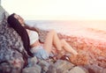 Dreamy lonely brunette hippie in the style of bokho chic sits on gray stones at the sea shore in the rays of the setting sun