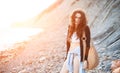 Dreamy lonely brunette hippie in the style of bokho chic sits on gray stones at the sea shore in the rays of the setting sun