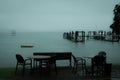 Dreamy photo of an empty beachside cafe table and chairs on a raint day. Small leasure boats anchored near the coast of