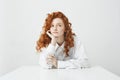 Dreamy tender young girl with red curly hair thinking dreaming sitting at table over white background. Royalty Free Stock Photo