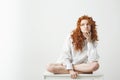 Dreamy tender young girl with red curly hair thinking dreaming sitting on table over white background. Royalty Free Stock Photo