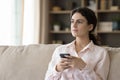 Dreamy thoughtful millennial girl holding smartphone, sitting on comfortable couch Royalty Free Stock Photo