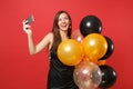 Dreamy woman in black dress celebrating holding air balloons doing taking selfie shot on mobile phone isolated on red Royalty Free Stock Photo