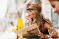 Dreamy woman in brown cap thinking about something, propping face with hand and holding newspaper. Outdoor portrait of Royalty Free Stock Photo