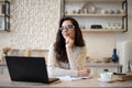 Dreamy woman sitting in kitchen at table with opened laptop, looking away and thinking, copy space Royalty Free Stock Photo