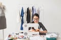 Dressmaker, tailor and fashion concept - Smiling female fashion designer using sewing machine and sitting behind her Royalty Free Stock Photo