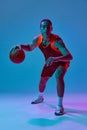 Dribbling ball . Dynamic image of teen boy in uniform, playing basketball against blue studio background in neon light