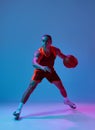 Dribbling ball . Dynamic image of teen boy in uniform, playing basketball against blue studio background in neon light