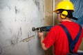Drilling a hole for the socket with a puncher. A young worker in a robe, a construction helmet and protective headphones with a Royalty Free Stock Photo