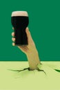 Drinking toast minimal party concept. Man hand holding pint of black stout beer pops out from pastel green floor