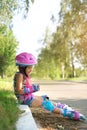 Droll girl in a helmet and roller skates sits on the curb of the sidewalk